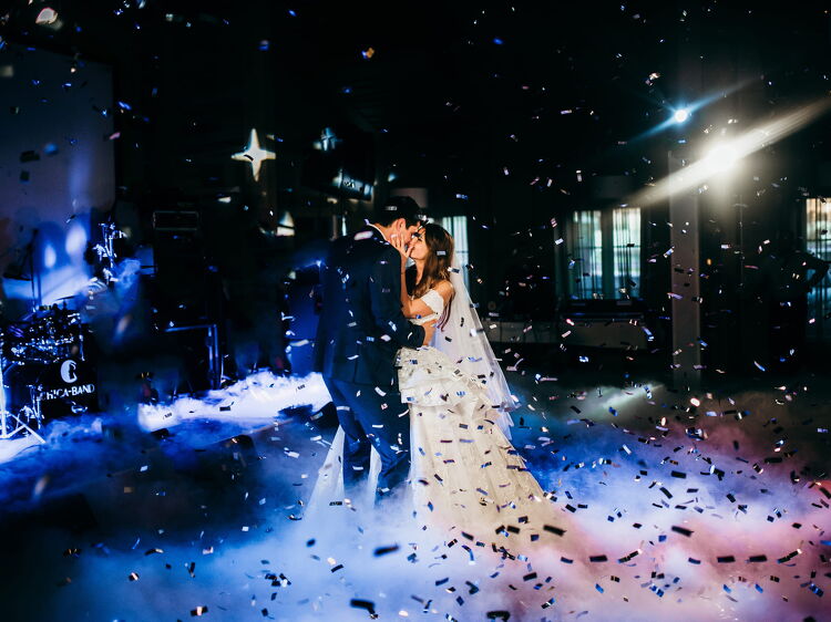 A bride and groom dancing under dramatic stage lighting with a smoky floor effect, surrounded by falling confetti during an evening wedding celebration.