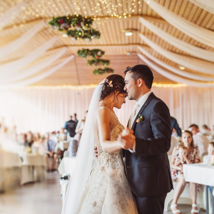 A bride and groom sharing their first dance in an elegantly decorated reception hall, with flowing fabric drapes and soft golden lighting in the background.