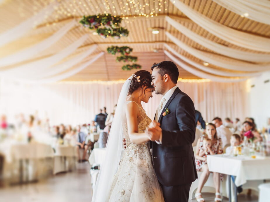 A bride and groom sharing their first dance in an elegantly decorated reception hall, with flowing fabric drapes and soft golden lighting in the background.