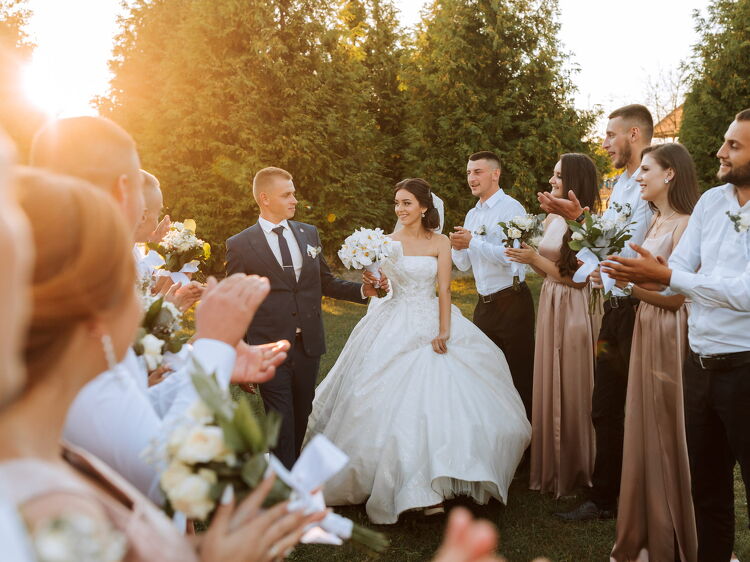 A wedding couple walking through an aisle of guests outdoors at sunset, with the guests clapping and holding bouquets, celebrating their special day.